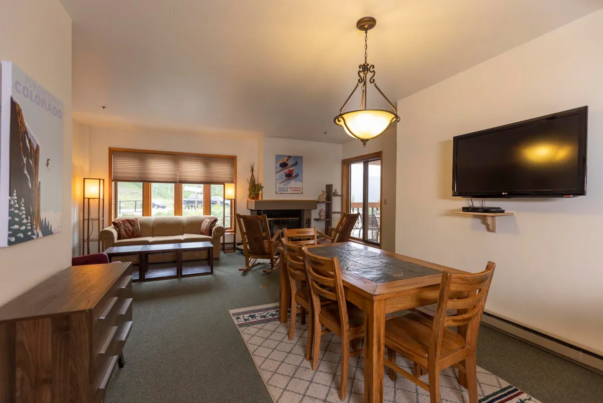 A spacious open-plan living area featuring a comfortable sofa and a wood-burning fireplace as the focal point. The scene includes a dining table in the background, a television for movie nights, and tasteful artwork, all bathed in warm, inviting lighting. Large windows fill the space with views of the alpine landscape, enhancing the sense of connection to the outdoors.