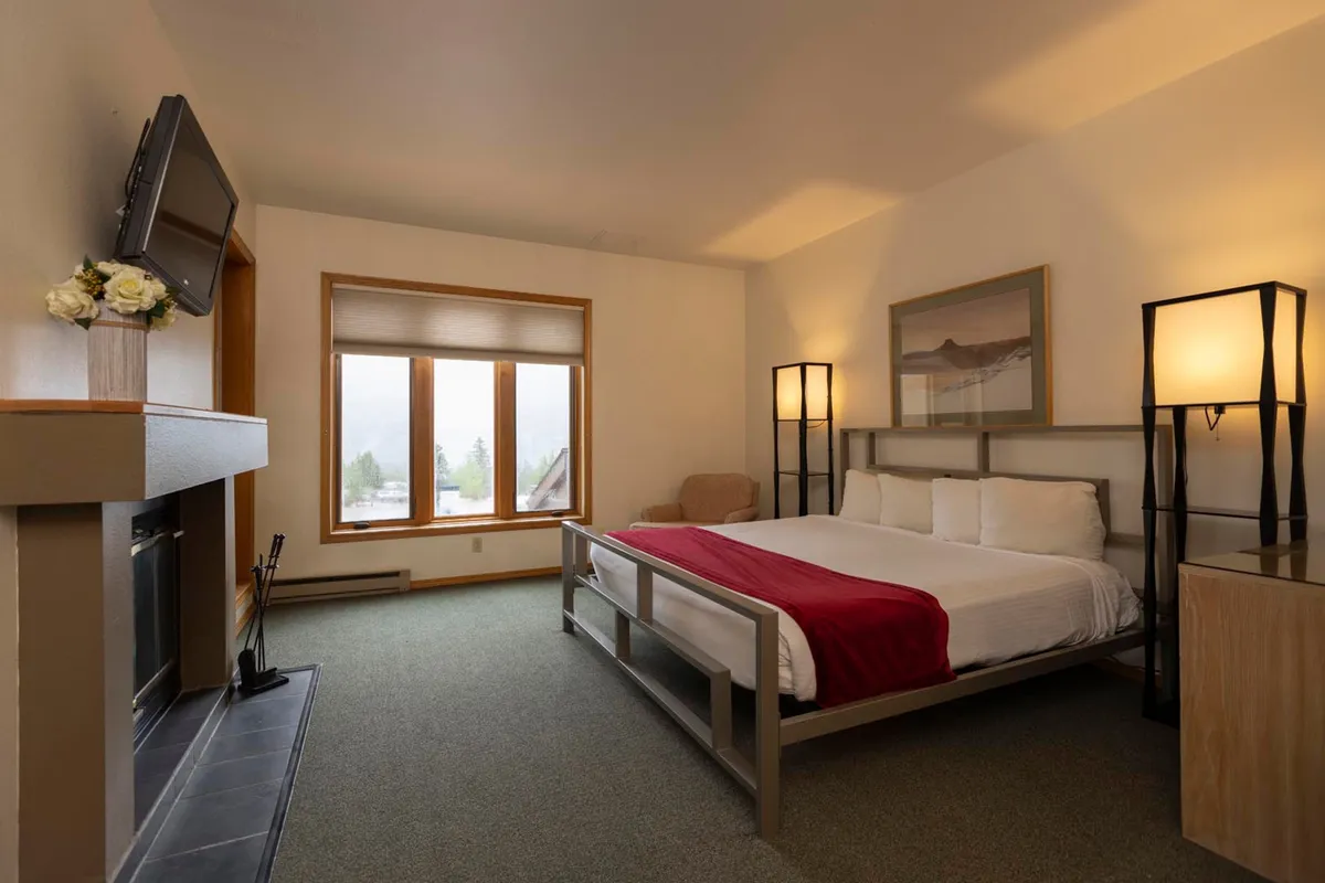 A serene bedroom featuring a plush bed with a bold red accent, large window letting in natural light, modern lighting fixtures, and a cozy fireplace. The overall styling is inviting and tranquil, suggesting both comfort and luxury. The spacious layout hints at ample storage options and a restful atmosphere.