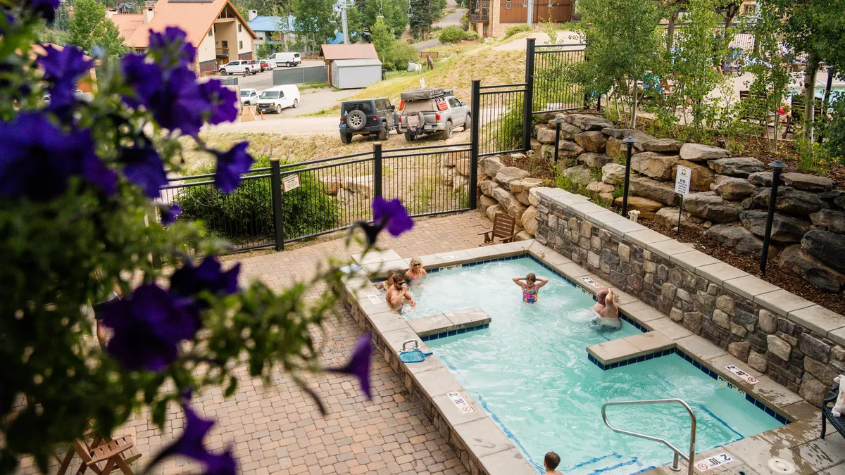 An outdoor heated pool surrounded by blooming flowers, with people joyfully swimming, sunbathing, and socializing. The poolside is lively and inviting, showcasing both relaxation and community in a scenic mountain setting.