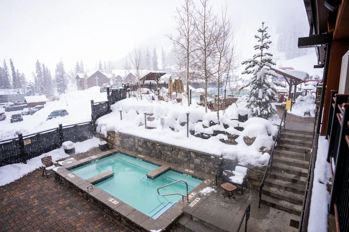This image features an outdoor hot tub set amid a snow-covered landscape, surrounded by trees and with steps leading down to the tub. The inviting steam and warm glow of the tub contrast beautifully with the serene winter environment, suggesting relaxation and luxury.