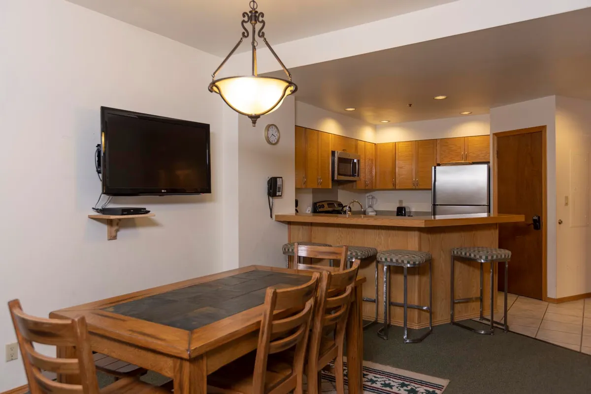 This image captures a cozy, well-lit dining area seamlessly connected to the kitchen counter, with a television visible in the background. The dining table is set for a meal, inviting conversation and togetherness, while the kitchen area is designed for easy meal preparation and social interaction.
