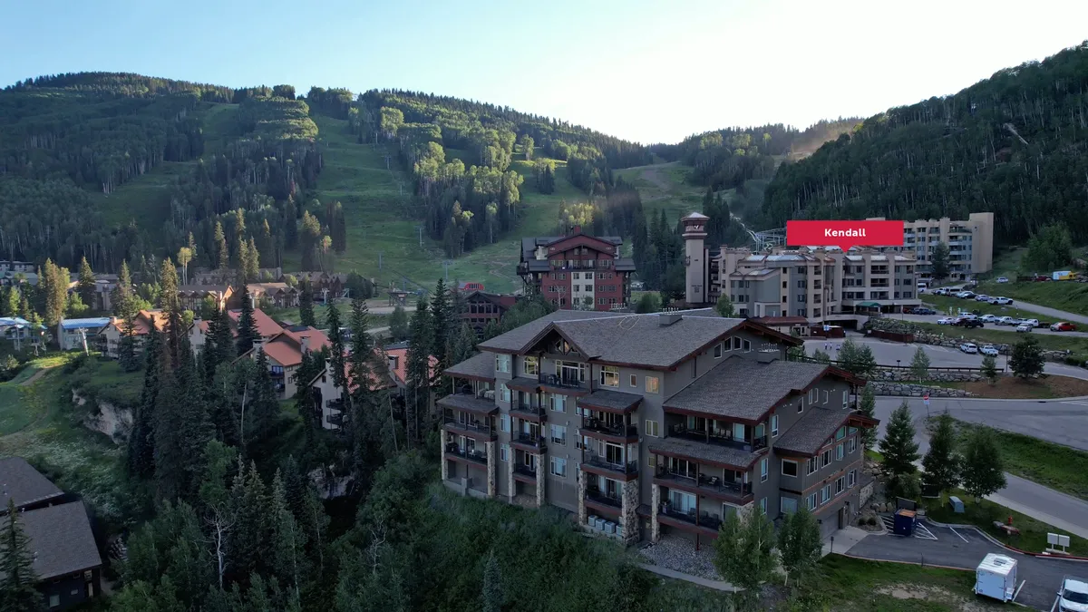 A wide exterior shot featuring lodge-style buildings set against a dramatic mountain backdrop, surrounded by lush greenery and a visible parking area. The setting invites thoughts of outdoor adventure and peaceful natural surroundings, with a clear sense of connection to the mountain environment.