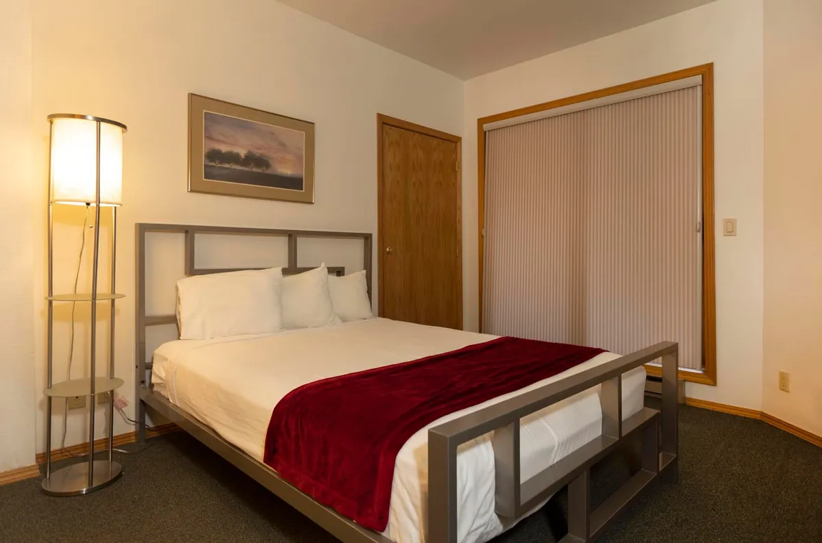 A cozy bedroom scene with a plush bed topped with a red throw blanket, complemented by a floor lamp, framed artwork, and a sliding door with blinds. The soft lighting and inviting decor create a restful nook ideal for reading, relaxing, or restorative sleep.