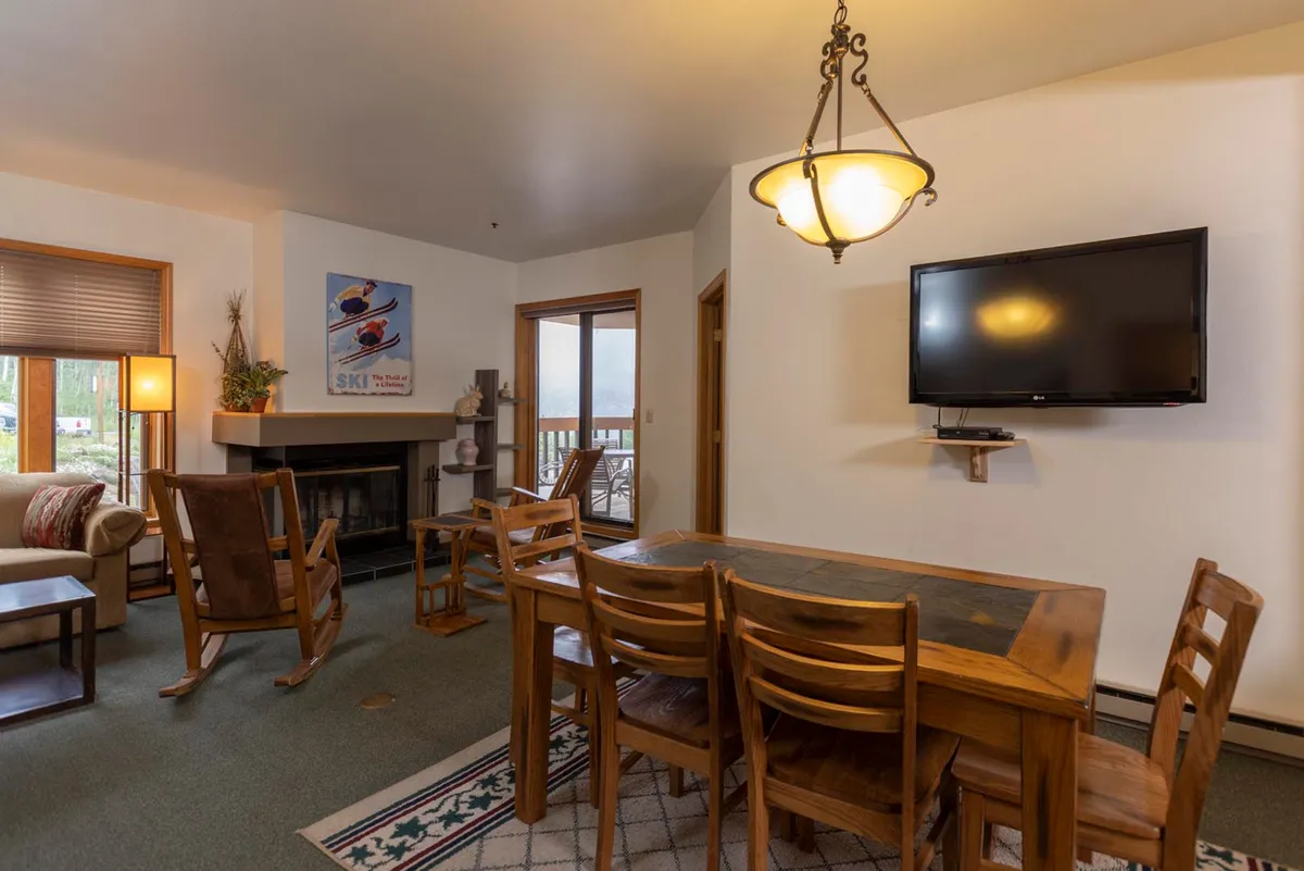A cozy interior scene with a dining table and comfortable chairs near a glowing fireplace. The arrangement invites group activities such as family dinners or game nights. The television and ambient lighting add to the space's versatility for entertainment and relaxation.