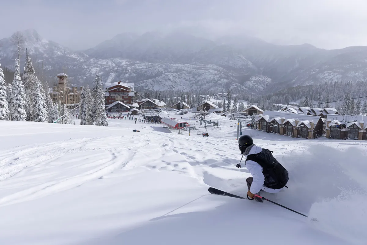 An action-filled exterior shot of a skier carving fresh tracks through pristine snow, set against a backdrop of snow-covered mountains and visible ski resort buildings. The lighting hints at late afternoon, adding warmth, while the energy of the skier conveys the peak of winter excitement.