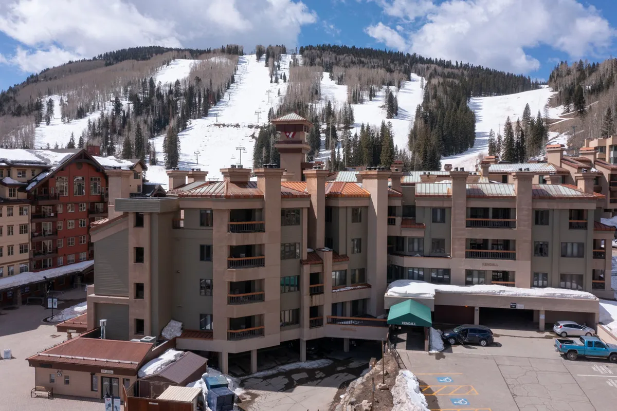 An inviting view of the resort building set against dramatic mountain slopes, with ski and snowboard tracks tracing the snow. The architecture blends seamlessly with the surrounding trees, while the parking area hints at convenient access for guests arriving for their mountain adventures.