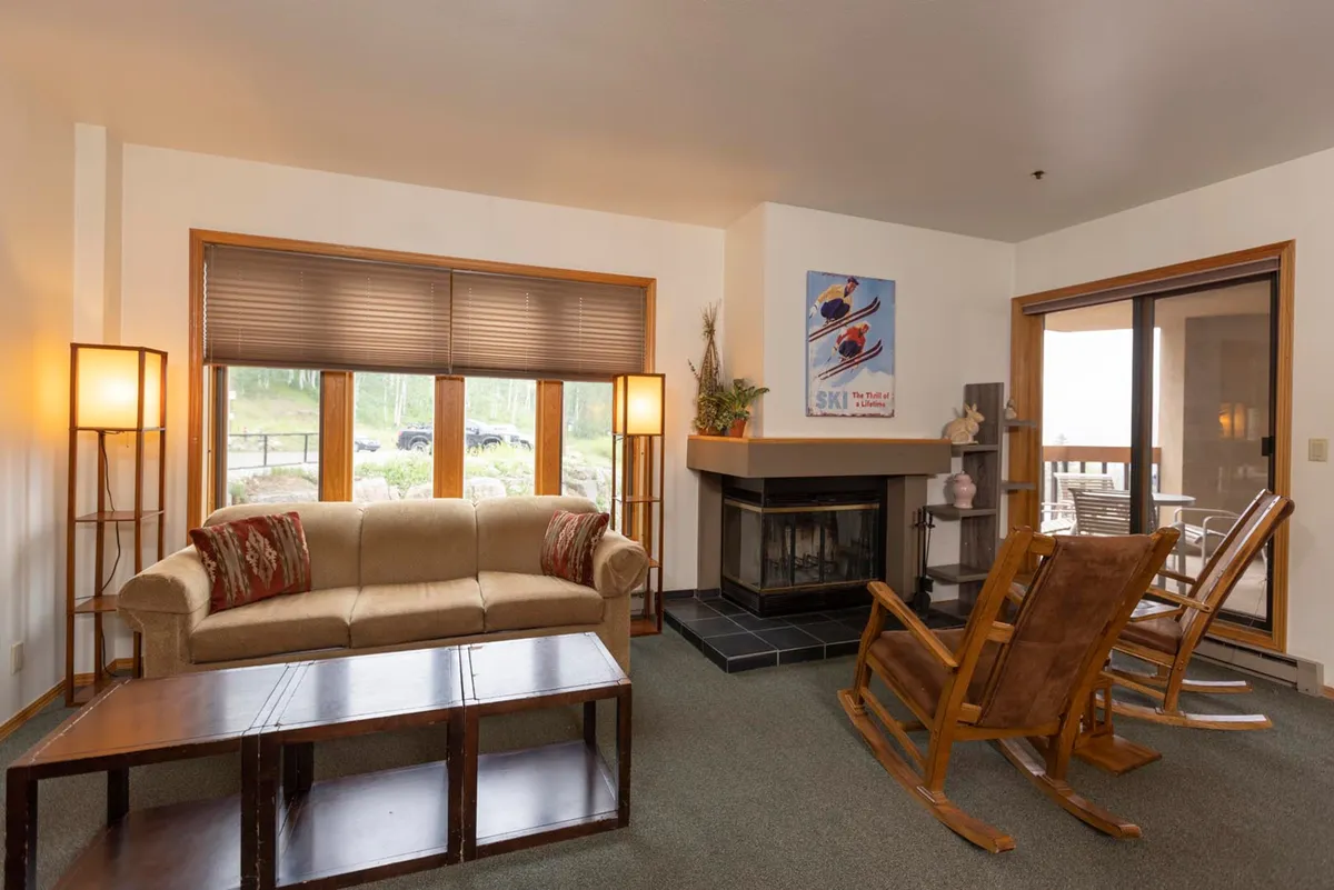 The image shows a warm, inviting living area with a plush sofa and cozy rocking chairs arranged around a wood-burning fireplace. Large windows let in natural light and offer glimpses of mountain scenery. The scene suggests a perfect setting for relaxation, reading, or gathering together after a day outdoors.