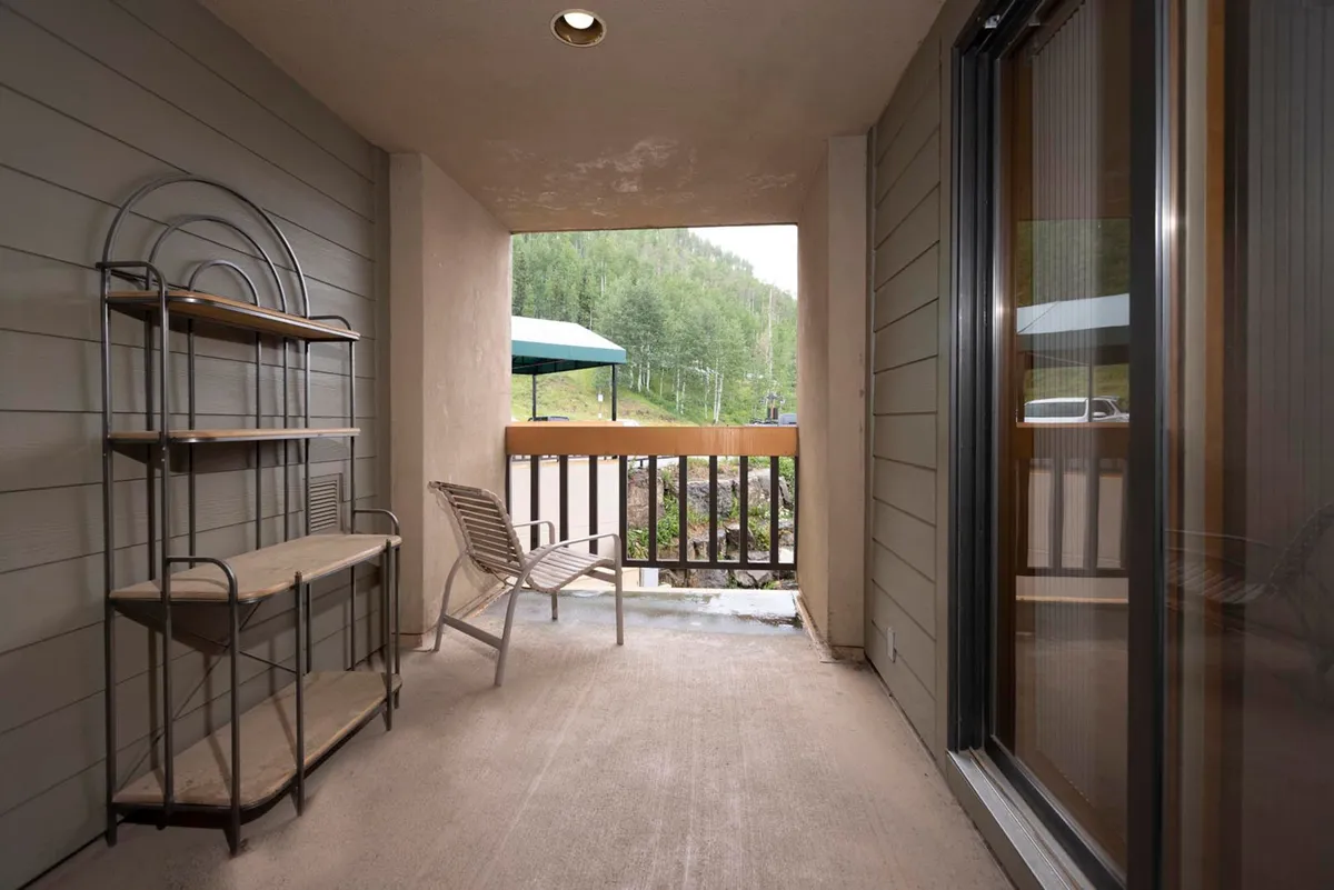 A cozy exterior balcony scene with a single chair and metal shelving unit, offering views through the balcony to the outdoors. The tranquil setup suggests a perfect spot to sip coffee, read, or enjoy the changing seasons while soaking in nature's sights and sounds.