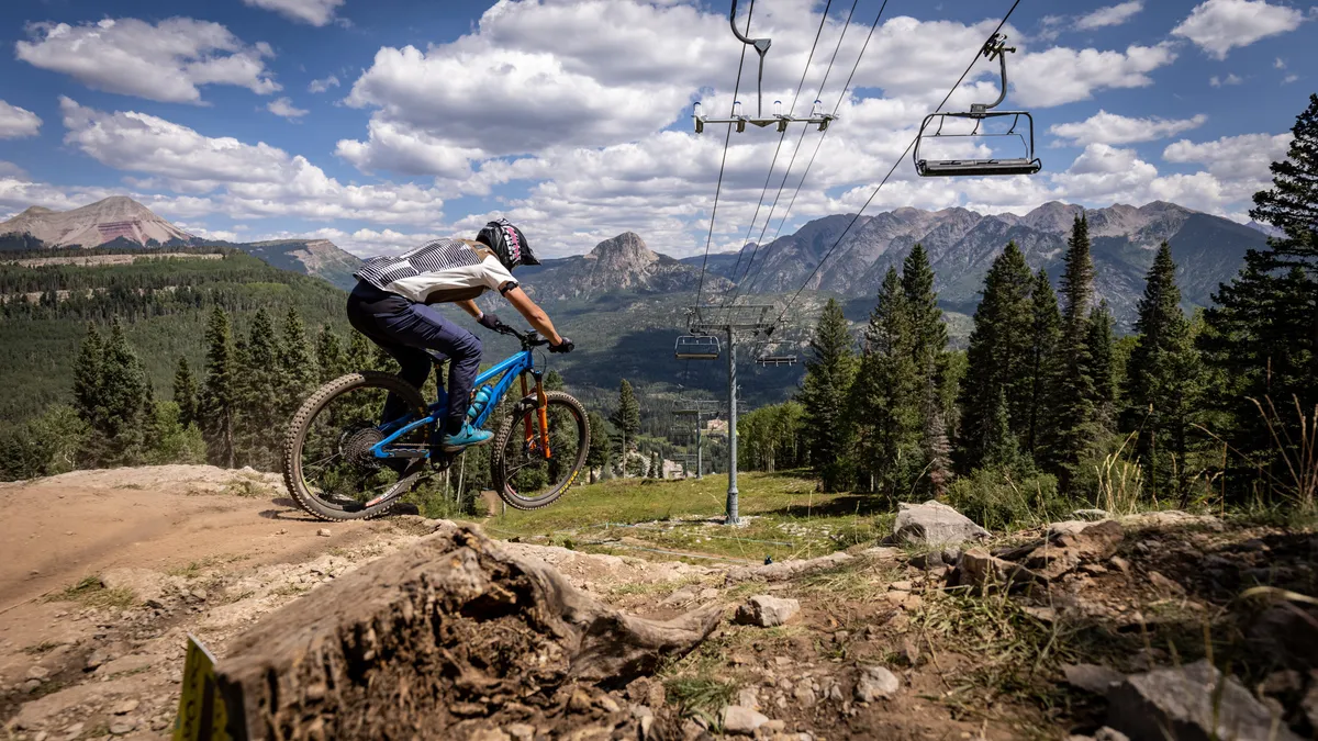 A dynamic exterior scene featuring a mountain biker in motion, a ski lift ascending the mountain, expansive mountain landscapes, and tall pine trees. The image perfectly captures both summer and winter adventure possibilities, with clear visual cues for mountain biking, hiking, and skiing. The energetic vibe communicates excitement and accessibility to outdoor recreation.
